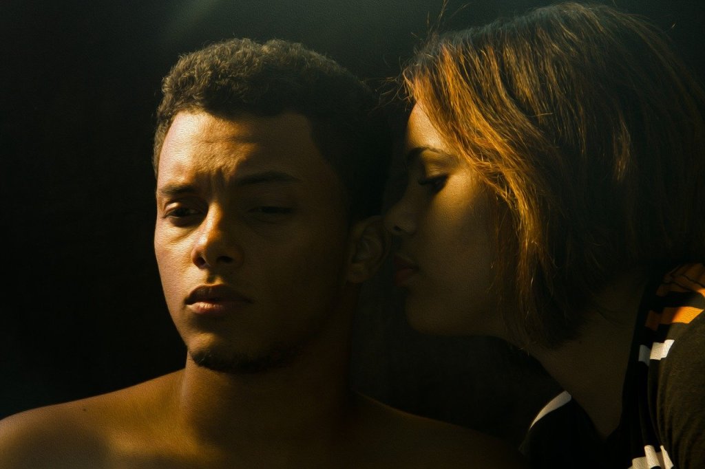 Image: young white person with chin-length blonde-brown hair whispering into the ear of an intense young brown person with short black hair.