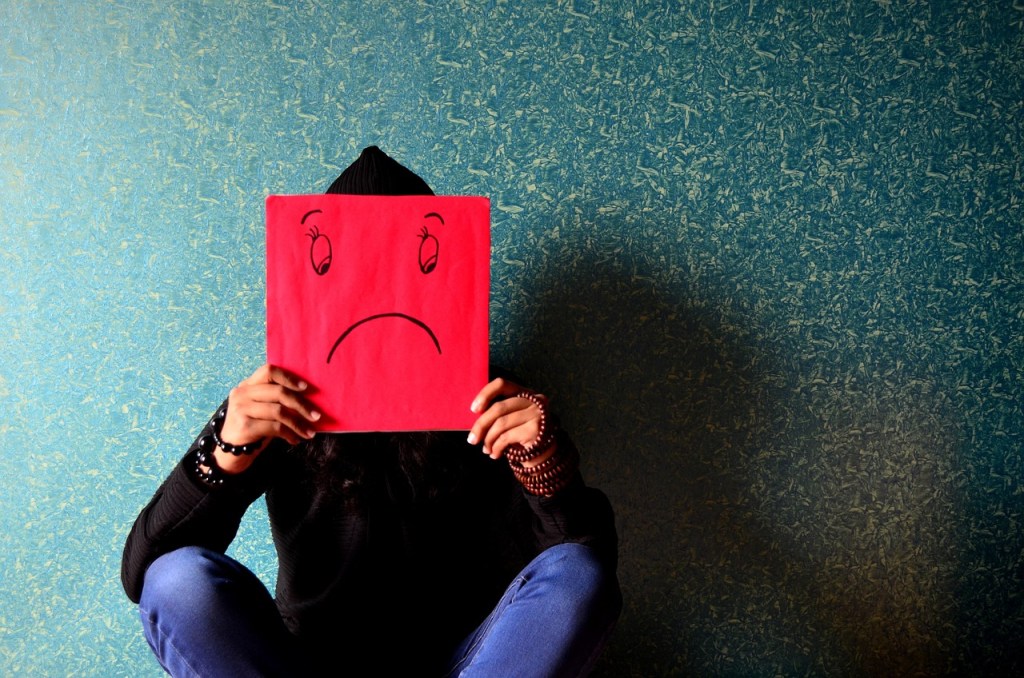 Image: seated person in hoodie and jeans holding a drawing of a sad face over their face